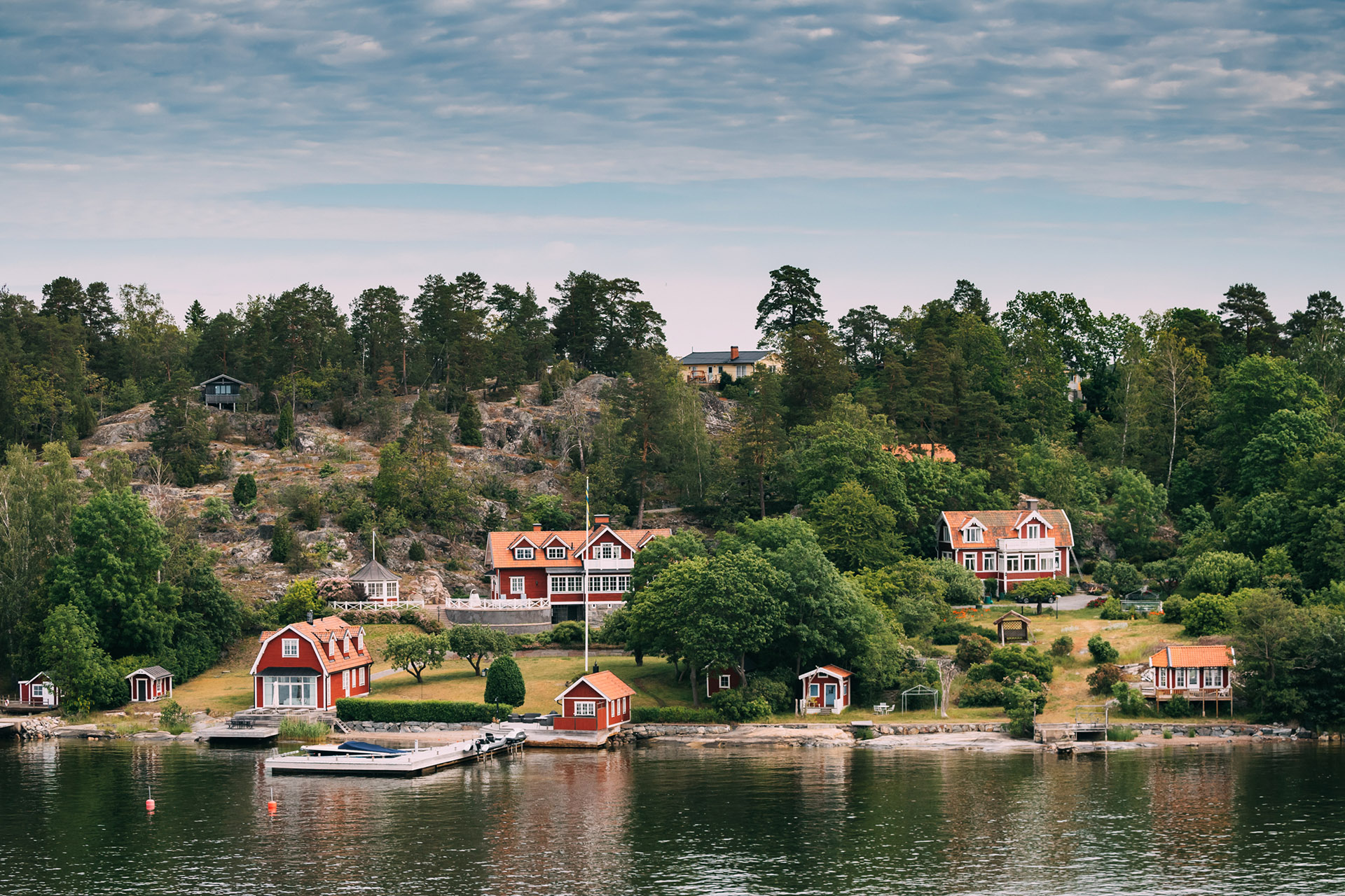 Sweden. Many Beautiful Red Swedish Wooden Log Cabins Houses On Rocky Island Coast In Summer Sunny Evening. Lake Or River Landscape.