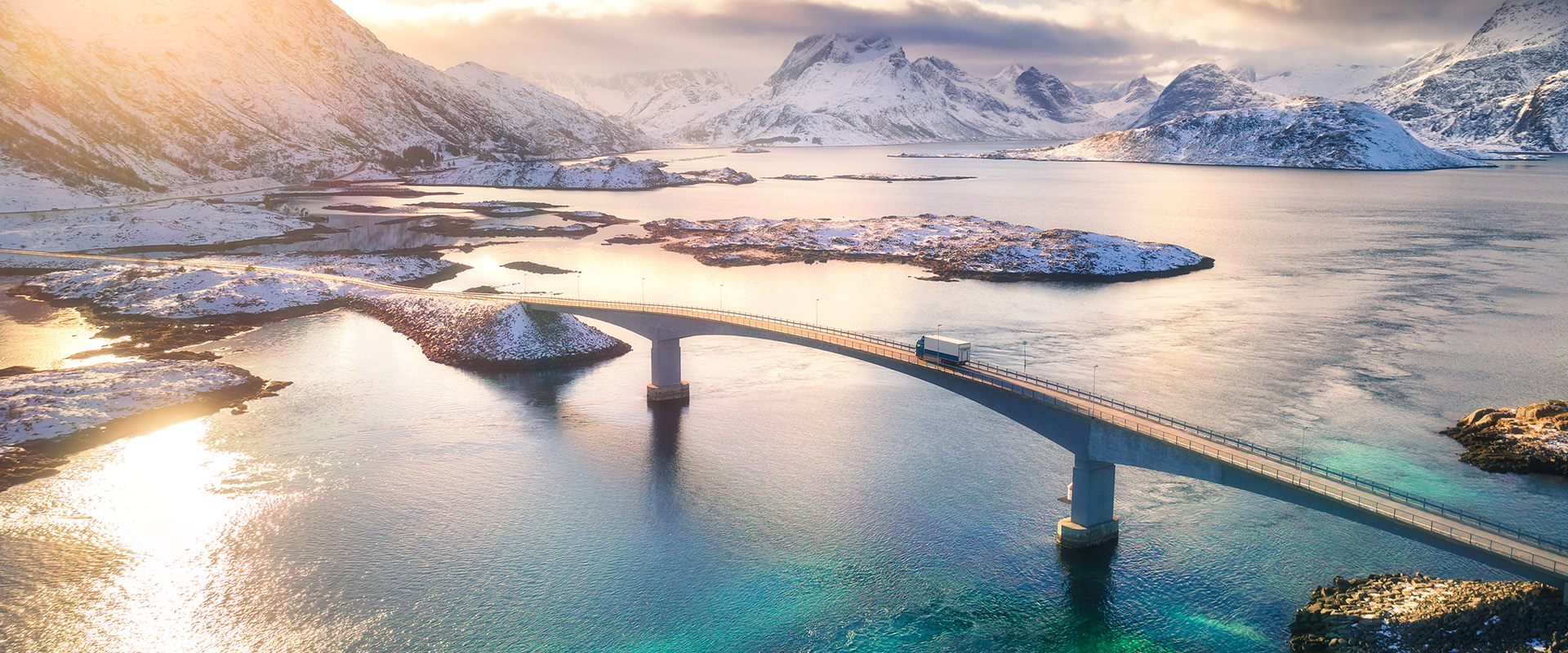 Aerial view of bridge over the sea and snowy mountains in Lofoten Islands, Norway. Fredvang bridges at sunset in winter. Landscape with blue water, rocks in snow, road and sky with cloads. Top view